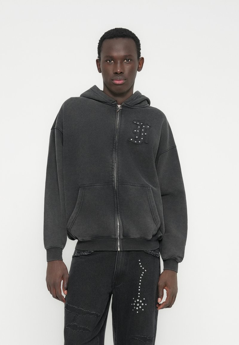 Grauer Kapuzenpullover mit Reißverschluss, Kapuze, großen Fronttaschen und dekorativen Nieten auf der Brust. Kombiniert mit schwarzen, verwaschenen Jeans mit Nietenakzenten.