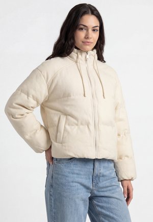 Frau trägt eine cremefarbene gesteppte Jacke mit Frontreißverschluss und Kordelzugkapuze, kombiniert mit hellblauen Jeans, steht mit einer Hand in der Hüfte.
