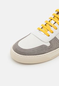 Grå semsket skinn og hvit lær sneakers med gule snører, med en avrundet tå og teksturert gummisåle med mønstret grep.