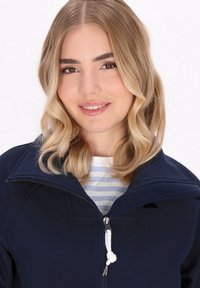 Tamsiai mėlynas softshell tipo džemperis su aukšta apykakle, užtrauktuku ir elastinga raišteliu. Jis turi jaukų vidinį pamušalą ir mažą logotipą ant krūtinės.