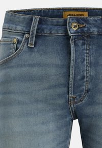 Blauwe spijkerbroek van denim met een knoop aan de voorkant, rits, riemlussen en een leren merklabel met de tekst "JACK&JONES" aan de binnenkant van de tailleband.