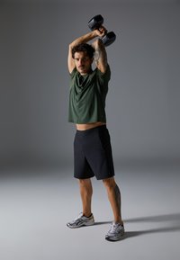 Green athletic shirt, black shorts, silver running shoes. Holds two black dumbbells overhead. Tattoo visible on right arm.