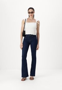 White smocked tank top with ties, paired with dark blue flared jeans and brown heeled sandals. Model holds a small black handbag.