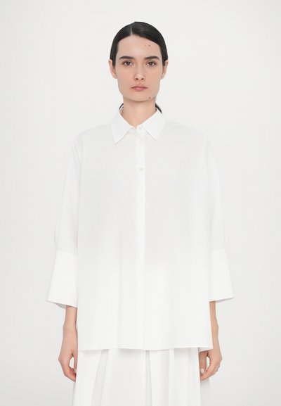 Chemise blanche à boutons avec un col pointu, coupe surdimensionnée et larges manches 3/4. Fabriquée en tissu lisse avec un léger éclat.