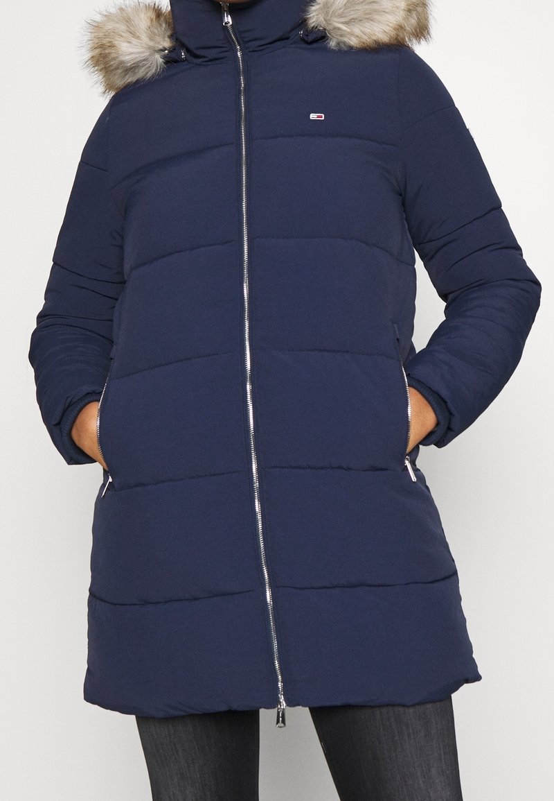 Marinblå pufferjacka med huva av konstpäls, horisontell sömnad, hel dragkedja och sidofickor. Har logotyp på bröstet.