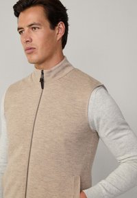 Gilet beige senza maniche con colletto alto, zip frontale e tasche laterali, indossato sopra una camicia a maniche lunghe grigia chiara. Texture liscia.