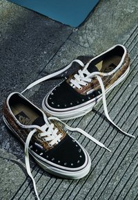 Pair of black and brown Vans sneakers with white laces and metal spikes on the toes resting on textured concrete pavement.