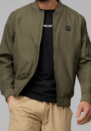 Homme portant une veste bomber vert olive non fermée sur un t-shirt noir avec un texte blanc, et un pantalon beige, une main dans la poche.