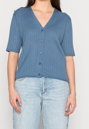 Kobieta w niebieskim prążkowanym rozpinanym sweterku z krótkim rękawem, zestawionym z jasnoniebieskimi, wysokim stanem jeansami na jednolitym tle.