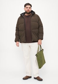 Polstret brun jakke med hætte, grå foring, beige bukser og brune sko. Grøn tote bag med dobbelte håndtag og lynlåslukning.