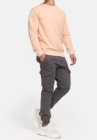 Lyse ferskenfarvet sweatshirt med lange ærmer, parret med mørkegrå cargo-bukser med flere lommer og hvide sportssko.