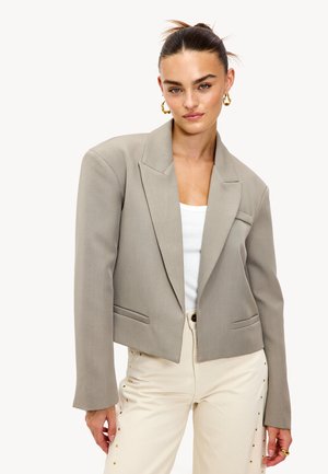 Blazer gris cropped avec revers, fabriqué en tissu lisse ; il présente deux poches avant, associé à un pantalon crème orné d'clous.