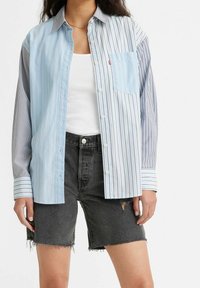 Camicia a righe blu e grigia con taschino, abbinata a pantaloni corti in denim neri con orli sfilacciati e dettagli distressed.