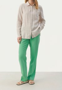 Lys beige linnedragt med knapper og lange ærmer, kombineret med grønne bukser med lige snit og hvide sandaler med et enkelt remdesign.