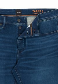 Mörkblå jeans med smal passform, knäppning med knappar, orange sömmar och synliga externa varumärkesetiketter "BOSS" och "TABER TAPERED FIT."