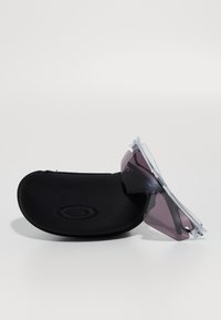 Sort hård solbrilleetui med Oakley-logo ved siden af wraparound sports solbriller med klar ramme og mørkt tonede linser.