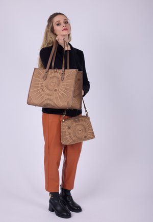 Frau in schwarzem Pullover und orangefarbenen Hosen, die zwei braune Handtaschen mit Sonnen- und Textdesigns hält, vor einem schlichten hellen Hintergrund stehend.