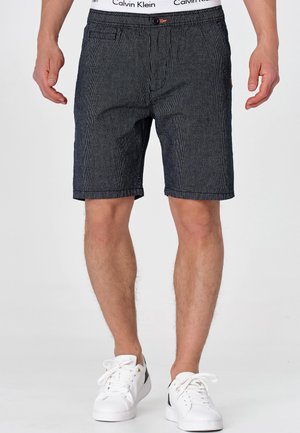 Mężczyzna ubrany w czarne teksturowane szorty z widocznym białym pasem Calvin Klein oraz białe trampki z ciemnym akcentem, stojący na białej podłodze.