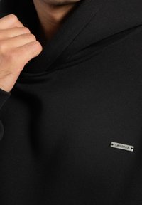 Zwarte hoodie gemaakt van zacht, gestructureerd stof met een hoge kraag en een metalen merkplaatje aan de voorkant, met een minimalistisch ontwerp.