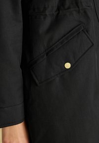 Schwarzer Stoffmantel mit einer auffälligen Brusttasche, ausgestattet mit einem goldenen Knopfverschluss und sauberer Nahtführung an den Kanten.