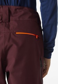 Bordeauxrode broek van gladde stof, met een oranje accent ritszak aan de achterkant en een zwarte ritszak aan de zijkant.