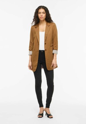 Kvinne som står iført brun blazer over hvit topp, sorte smale jeans og svarte åpne høyhælte sko mot hvit bakgrunn.