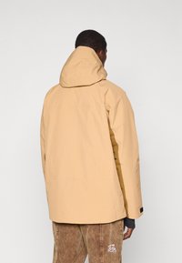 Άνδρας με μπεζ ζακέτα με κουκούλα και καφέ παντελόνι από βελουτέ στέκεται με την πλάτη προς την κάμερα σε απλό φόντο.