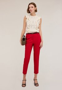 Witte gebreide top met gerafelde randen, voorzien van een gestructureerd patroon, gecombineerd met op maat gemaakte rode broeken en zwarte sandalen met bandjes.