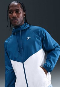 Homme aux cheveux tressés portant une veste coupe-vent Nike bleue et blanche, debout avec une main dans la poche devant un fond gris foncé.