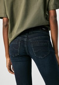 En person som bär mörkblå jeans med broderad vågmönster på bakfickorna och en olivgrön lös t-shirt.