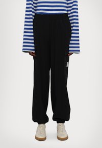 Μαύρο παντελόνι με φαρδιά μπατέτα και ελαστική μέση, με πλαϊνές τσέπες και ένα μικρό λογότυπο. Φοριέται με μπλούζα μακρυμάνικη σε μπλε και γκρι ρίγες.