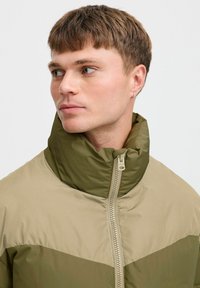 Chaqueta acolchada con un alto cuello, paneles en color beige y verde oliva, y un cierre frontal con tirador metálico.