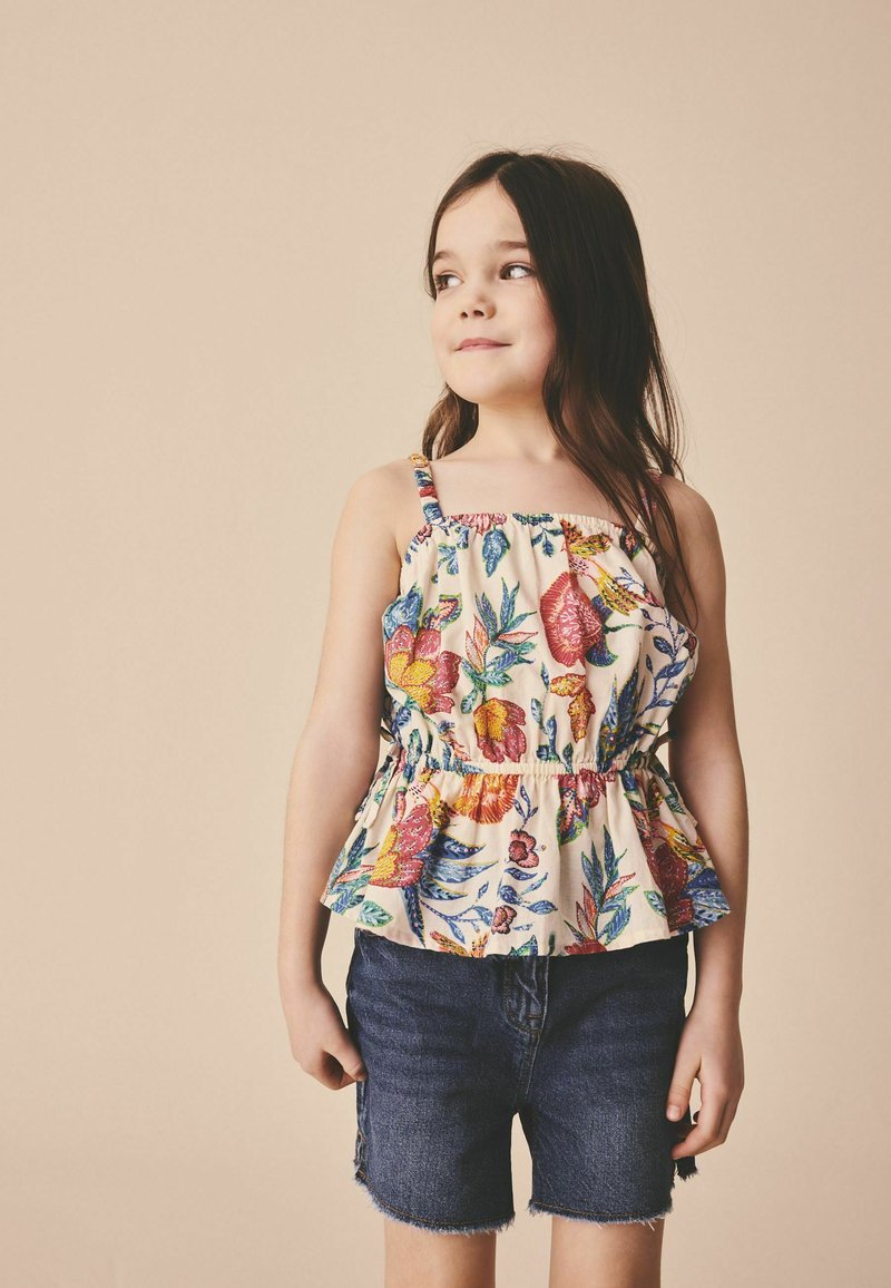 Niña joven con una blusa sin mangas colorida con flores y shorts de mezclilla oscuros, de pie y mirando hacia un lado contra un fondo beige.
