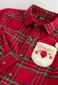 Rotes Karomusterhemd mit braunen Knöpfen, das auf der Brusttasche einen kleinen Aufnäher mit dem Gesicht eines weißen und roten Weihnachtsmanns zeigt.