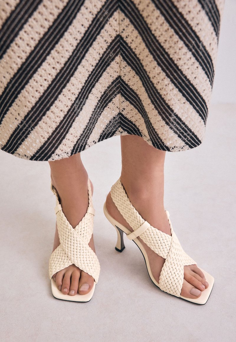 Sandales à talons hauts tissées couleur crème avec un design à bout carré et des brides croisées. Le matériau semble texturé, adapté à diverses occasions.