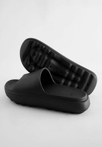 Sorte slidesandaler lavet af gummi med en tekstureret fodseng og en glat rem på toppen. Sålens underside har et mønstret design med drænhuller.