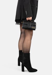Bottes noires à talon stiletto, en tissu texturé avec des accents scintillants. Portées avec une mini-jupe noire, des collants transparents et un sac à main noir.