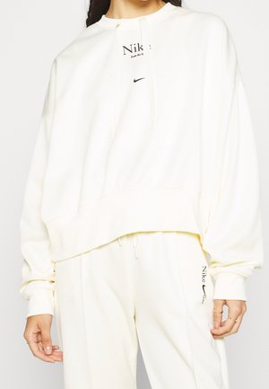 Osoba nosząca luźną białą bluzę Nike i dopasowane białe spodnie dresowe z czarnymi logo Nike na piersi i lewym udzie.