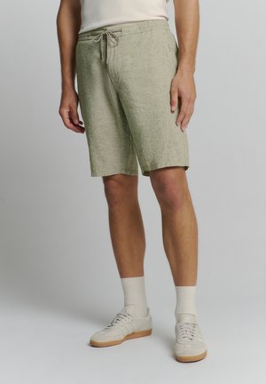 Shorts vert clair, texturés, avec une taille élastique et un cordon de serrage, offrant une coupe relaxée et une longueur jusqu'au genou. Associés à des chaussures et des chaussettes crème.