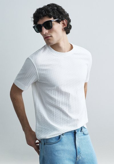 Camiseta blanca texturizada con un sutil patrón geométrico, combinada con jeans azul claro. La modelo lleva gafas de sol negras y tiene el cabello rizado.