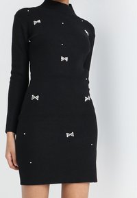 Robe noire côtelée à manches longues, col haut, ornée de petites perles en forme de perles et d'accents en forme de nœud sur le tissu.