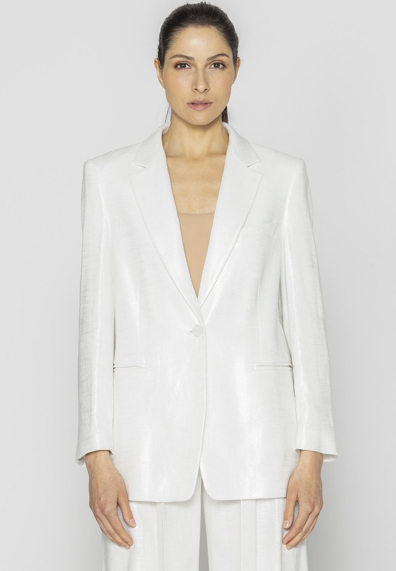 Biała marynarka skrojona z błyszczącego, teksturowanego materiału. Posiada wyłogi w szpic, zapięcie na jeden guzik oraz boczne kieszenie. Czysta, strukturalna sylwetka.