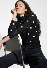 Zwarte lange mouwen top met grote crèmekleurige stippen, een hoge hals en een slanke pasvorm, gecombineerd met donkere spijkerbroek, zittend op een metalen stoel.