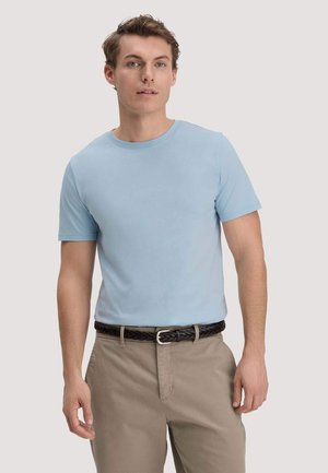 Jonge man die een lichtblauw T-shirt met korte mouwen, beige broek en een donker gevlochten riem draagt, staand tegen een effen achtergrond.