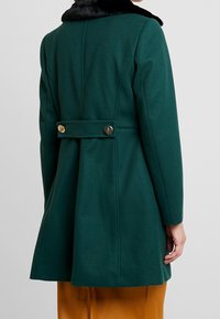 Cappotto verde in lana con colletto di pelliccia sintetica nera, forma strutturata, dettagli sul retro e due bottoni metallici in vita.
