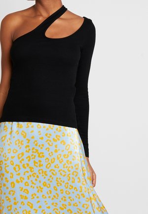 Frau trägt ein schwarzes Oberteil mit einem Ärmel und einem Cut-out an der Schulter sowie einen hellblauen Rock mit gelbem Leopardenmuster.