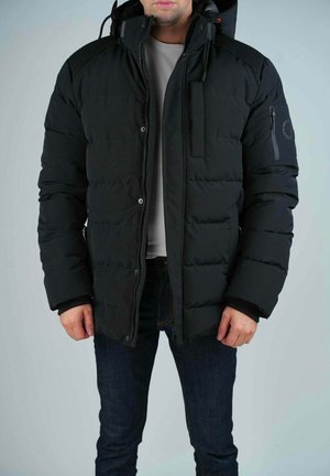 Homme portant une veste d'hiver matelassée noire avec capuche, fermeture éclair et boutons-pression, associée à un jean foncé et une chemise gris clair.