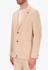 Blazer beige su misura in un tessuto testurizzato, con due bottoni, tasche a toppa e un piccolo distintivo multicolore sul rever.