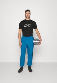 Camiseta negra de Nike con logo, pantalones deportivos azules con franjas laterales, zapatos de fútbol negros, sosteniendo un balón de fútbol gris y blanco.