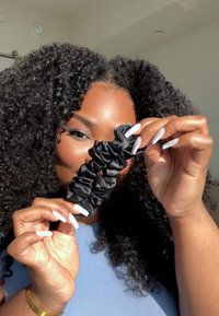 Svart satenghårstrikk med en rynket design, holdt mellom fingrene. Lange, hvite manikyrte negler er synlige. Bakgrunnen er svakt uskarp.